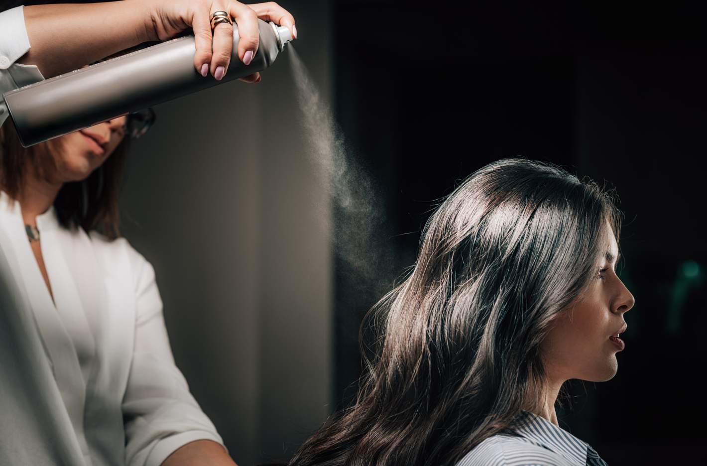 A hair stylist spraying hair spray on a seated woman's long black hair. Concept of uisng hair styling products along with keratin hair building fibers.